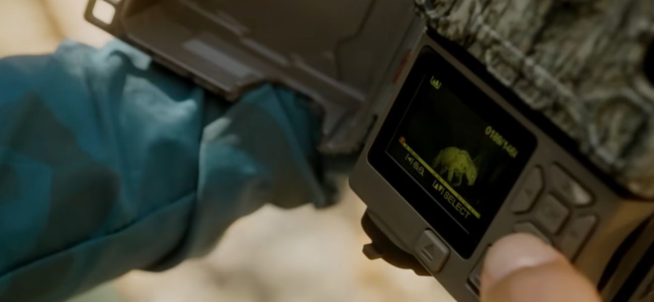 Close-up de um dispositivo eletrônico portátil, segurado por uma pessoa com luvas, mostrando na tela a imagem de um animal semelhante a uma onça em visualização térmica, em um ambiente externo.