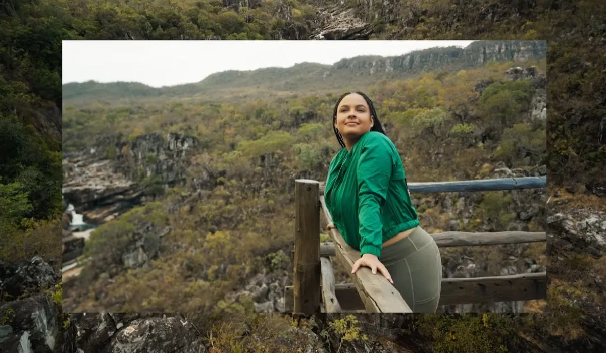 Uma mulher sorridente com uma jaqueta verde e calças de ginástica verde-oliva está encostada em uma cerca de madeira, olhando para a câmera com uma paisagem montanhosa e verde ao fundo.