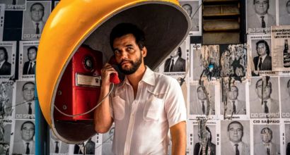 Homem usando telefone público vermelho sob cobertura amarela, em frente a parede com cartazes antigos em preto e branco.