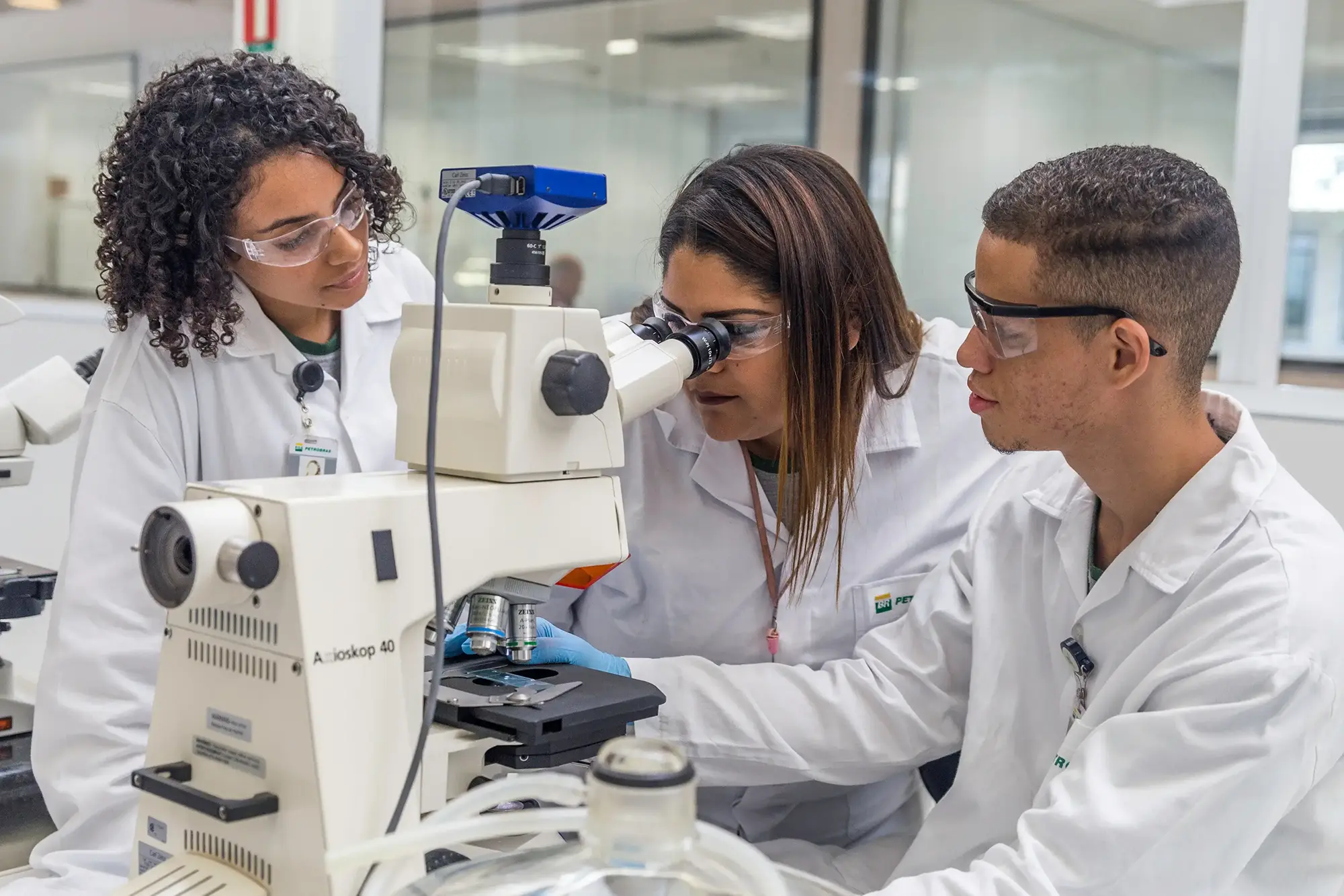 Mulher utilizando microscópio. Ao seu lado, outros dois cientistas conversando com ela.