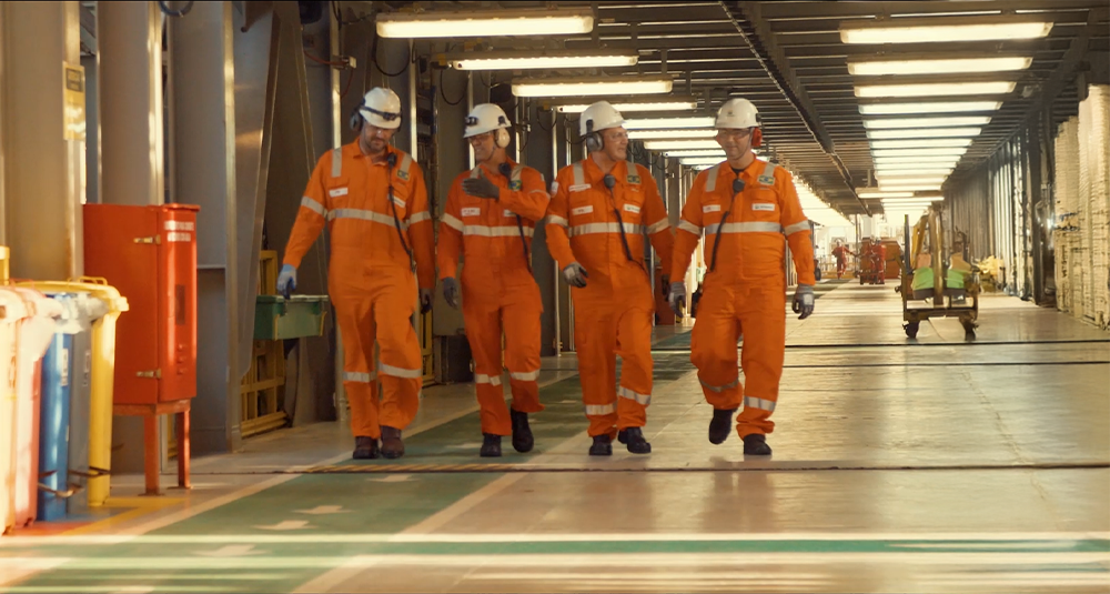 Quatro pessoas com uniforme laranja da Petrobras caminhando em um corredor.
