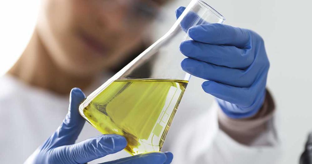 Mão com luva azul segurando um vidro de laboratório com óleo claro no interior.