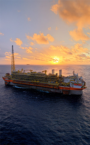 Plataforma FPSO Alexandre de Gusmão no mar durante o pôr do sol, com estrutura industrial destacada e embarcações ao fundo.