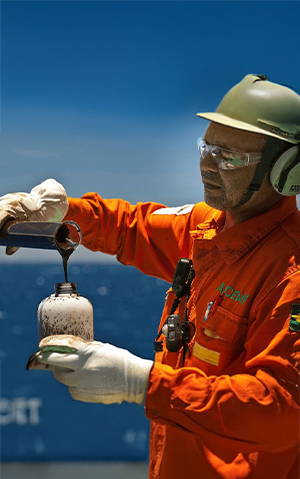 Homem com uniforme da Petrobras e proveta de petróleo com o mar ao fundo.