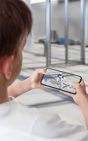 Um menino com celular em mãos, utilizando um aplicativo de realidade aumentada para visualizar um robô em sua frente.
