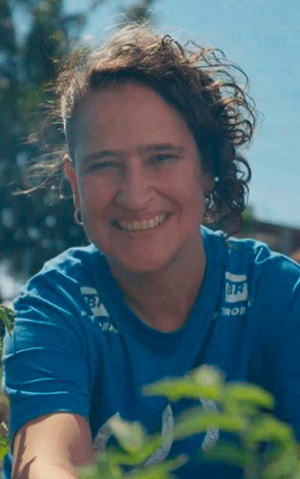 Mulher, com camisa do Projeto Corredor Caipira, sorri para a câmera em meio às folhagens de espécie nativa da Mata Atlântica, replantada pelo Projeto Corredor Caipira.