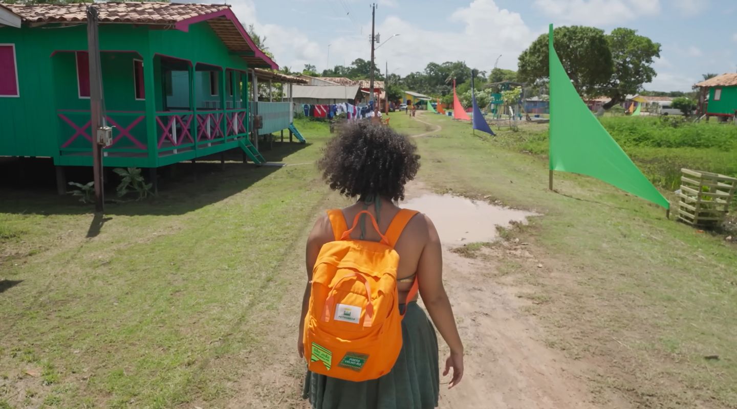 Pessoa caminhando em uma vila com casas coloridas, carregando uma mochila laranja sob céu ensolarado.