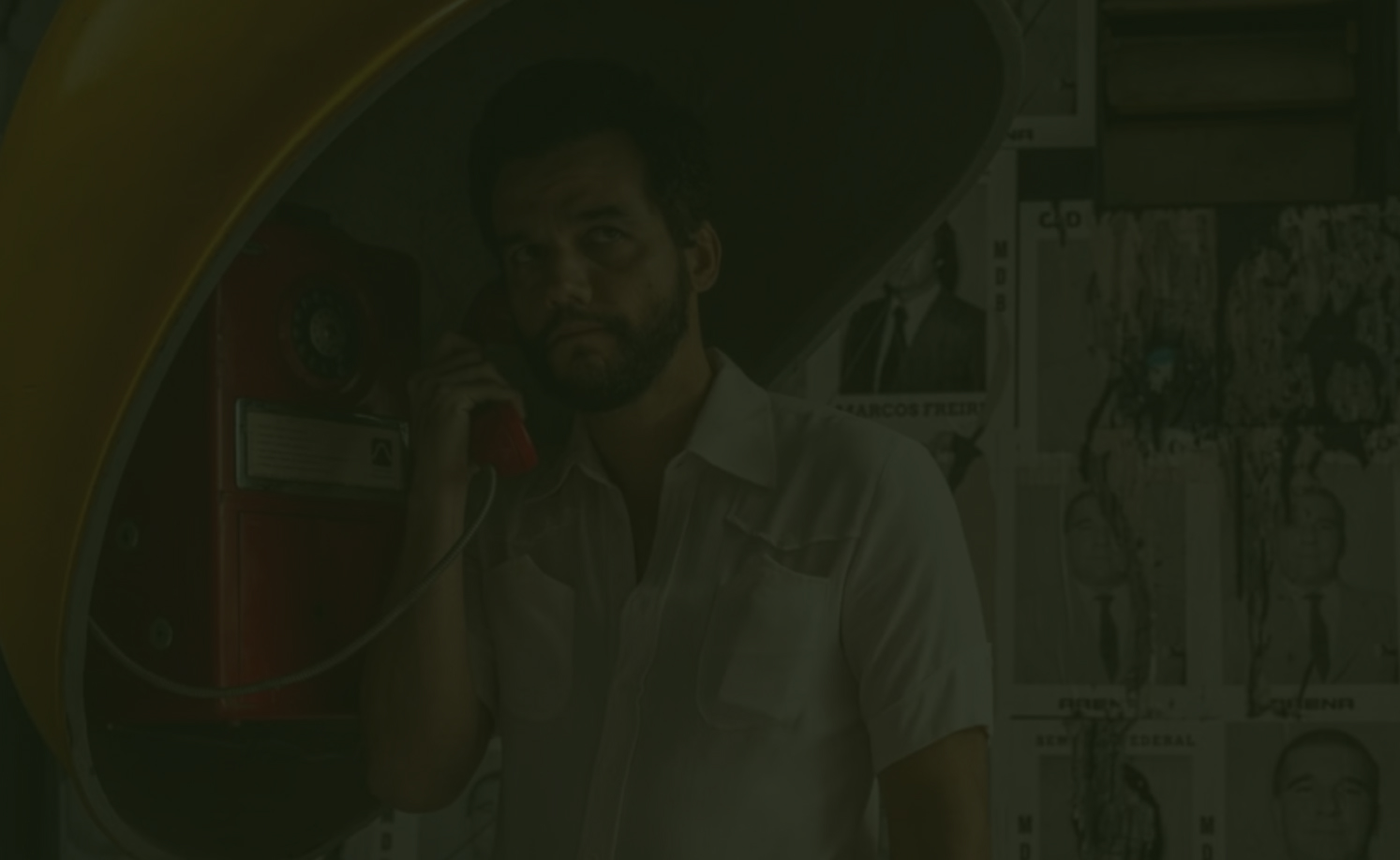 Homem de barba usa telefone público antigo em frente a cartazes políticos.