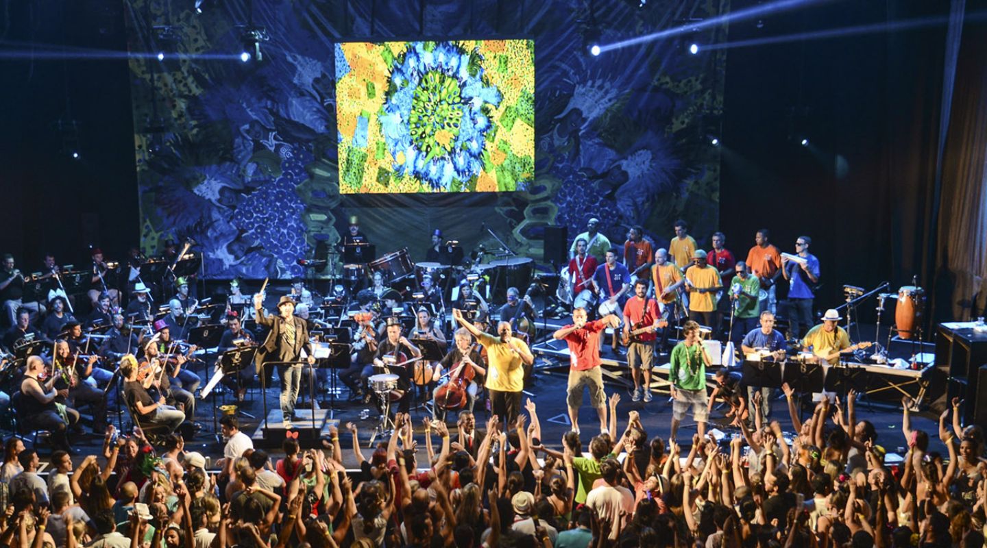 Apresentação musical em grande palco com orquestra, artistas e público celebrando um festival cultural.
