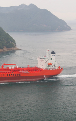 Navio petroleiro de casco laranja e vermelho navegando em águas calmas, contornando uma ilha montanhosa e arborizada.
