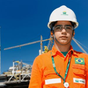 Jovem trabalhador da Petrobras em destaque, usando uniforme laranja, capacete branco e óculos de proteção, com uma plataforma de petróleo ao fundo sob céu azul.