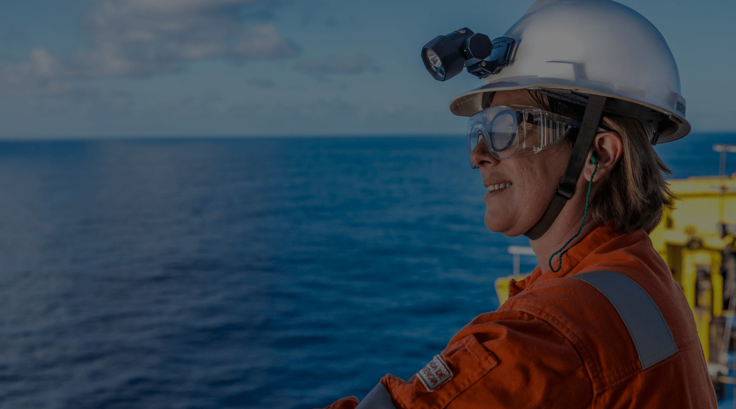 Profissional com capacete e óculos de proteção em plataforma offshore, observando o mar ao fundo.
