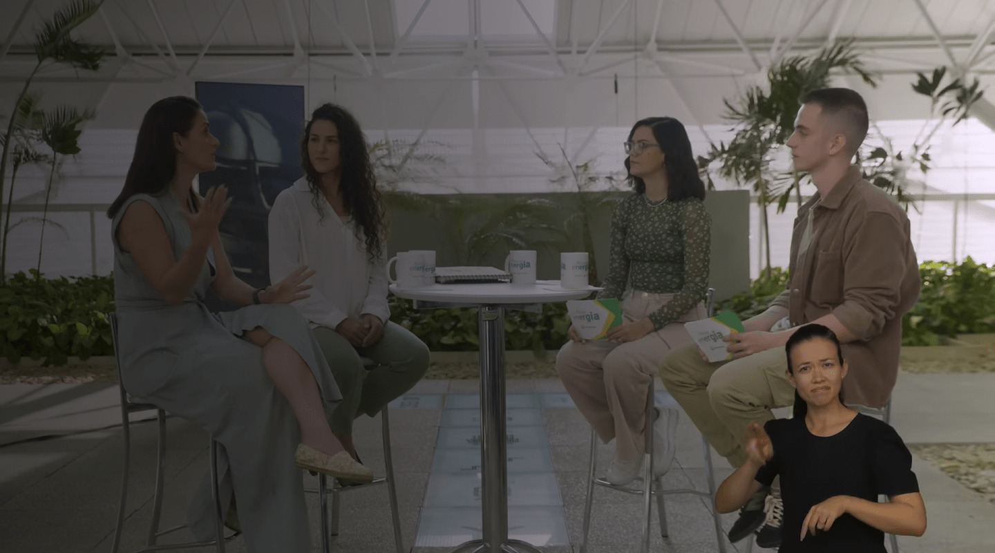 Grupo de pessoas sentadas em roda conversando em ambiente interno com plantas, com intérprete de Libras no canto da imagem.
