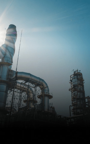 Refinaria com torres e dutos industriais sob céu azul, iluminada pelo sol.