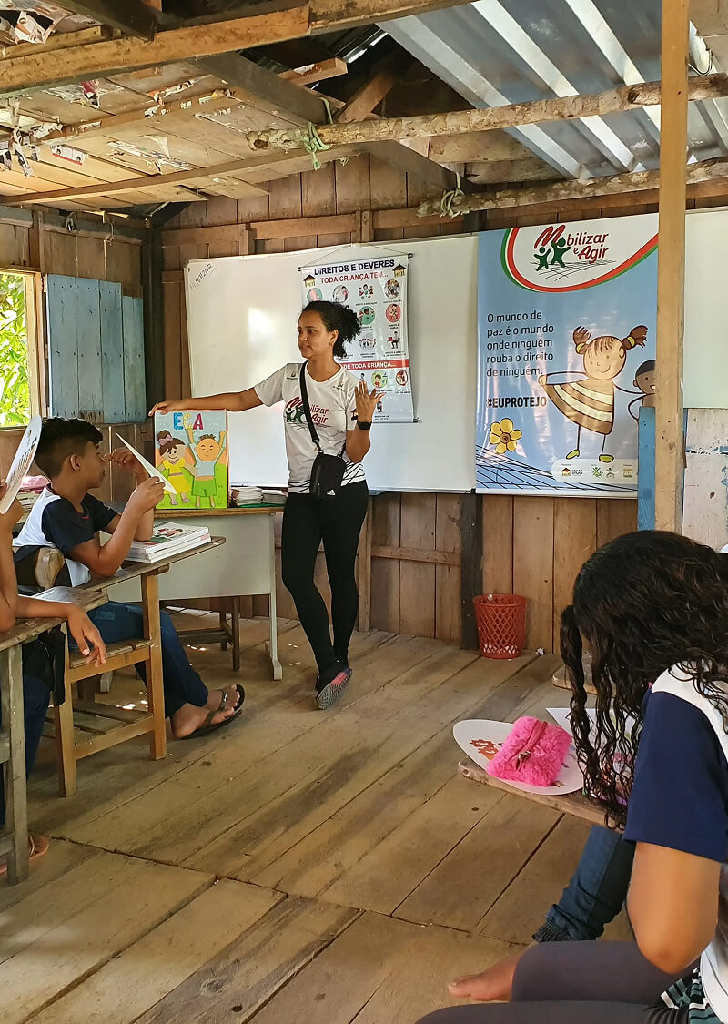 Professora mulher fala com crianças sentadas em uma sala de aula com paredes de madeira.