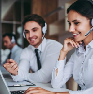 Fotografia de um homem e uma mulher, ambos usando camisa social, sentados em frente a um computador com fone de ouvido com microfone.