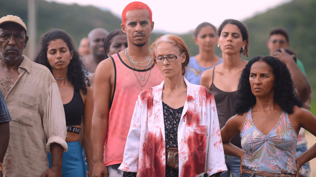 Atriz Sônia Braga com jaleco branco manchado, em frente a mais de dez outras pessoas, em cena do filme Bacurau.