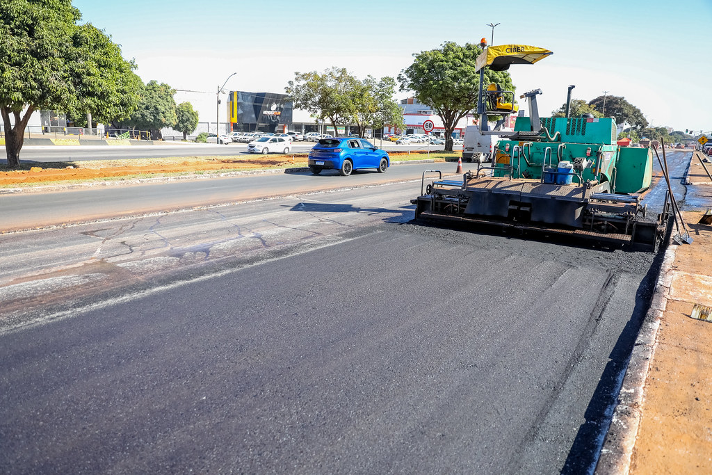 Máquina pavimentadora aplicando asfalto em via urbana, com carros circulando ao fundo.