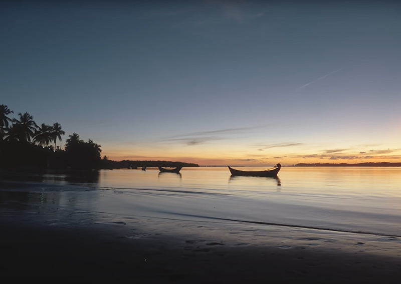 Pôr do sol na praia, com canoas flutuando e coqueiral na orla.