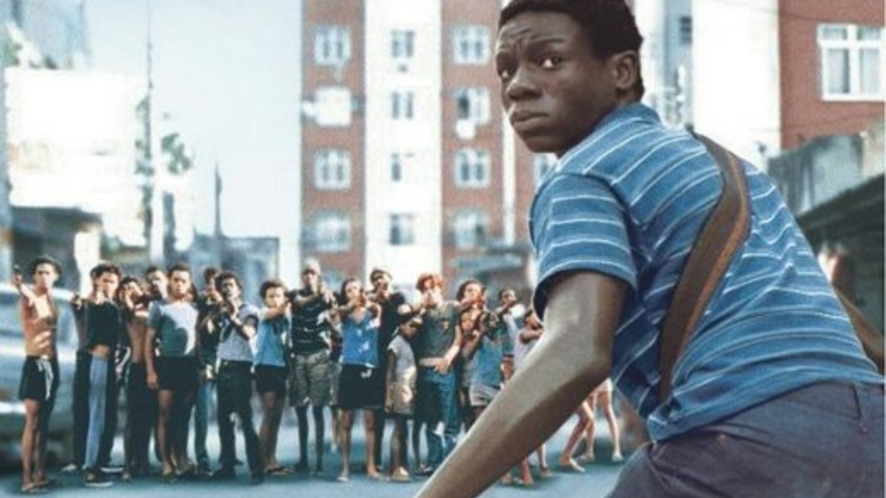 Jovem olha para trás com expressão tensa enquanto um grupo de pessoas ao fundo aponta armas em sua direção em uma rua urbana.