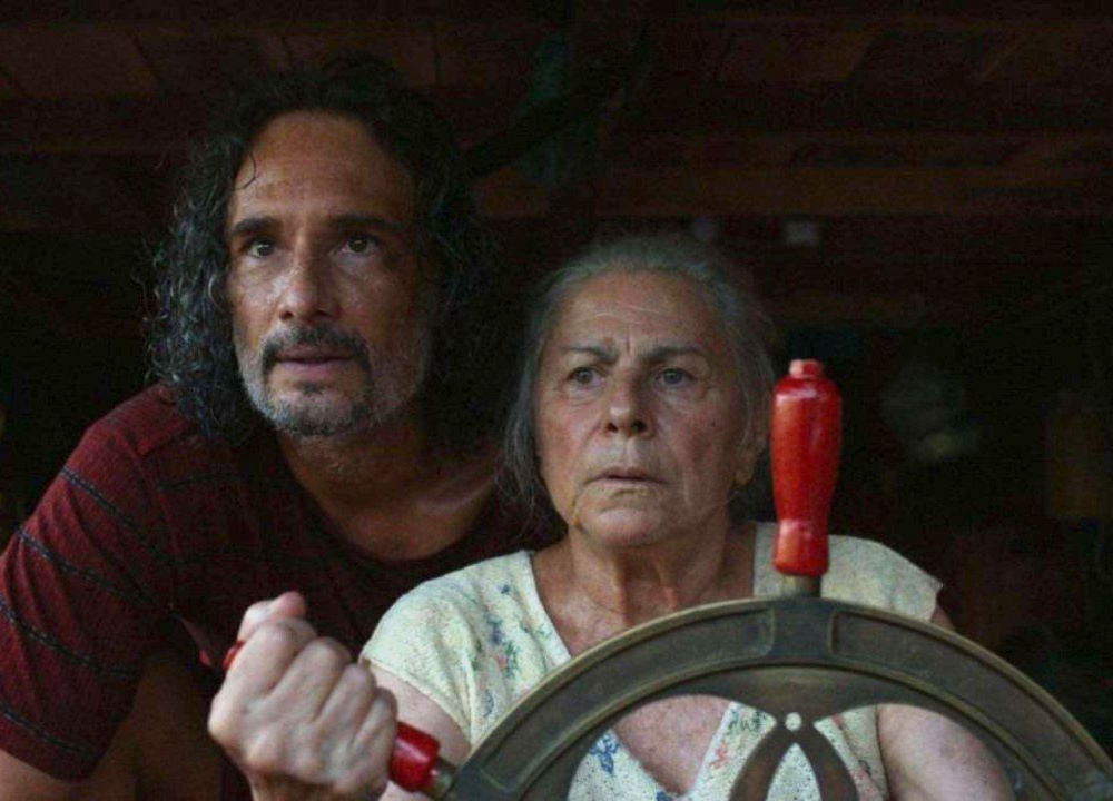 Homem e mulher idosa seguram o leme de uma embarcação e observam algo à frente com expressão de tensão.