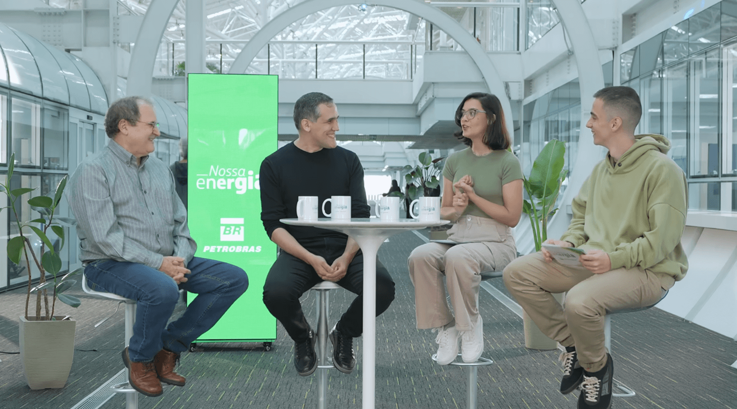 Uma mulher e três homens em ambiente corporativo moderno, sentados em bancos altos ao redor de uma mesa com canecas, com um painel verde da Petrobras ao fundo exibindo a frase "Nossa energia".