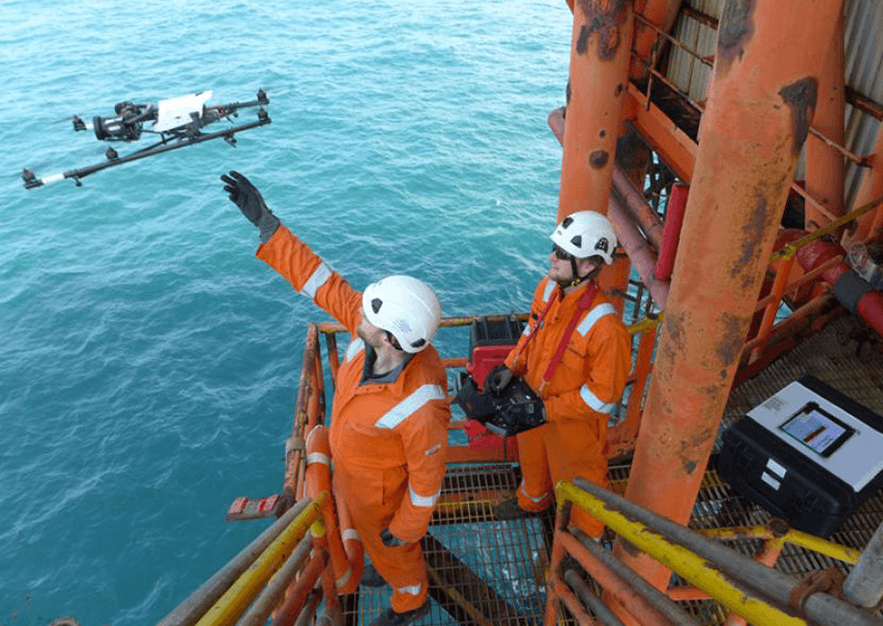 Dois trabalhadores em uma plataforma offshore operam um drone sobre o mar, utilizando equipamentos de segurança como capacetes e macacões laranja.