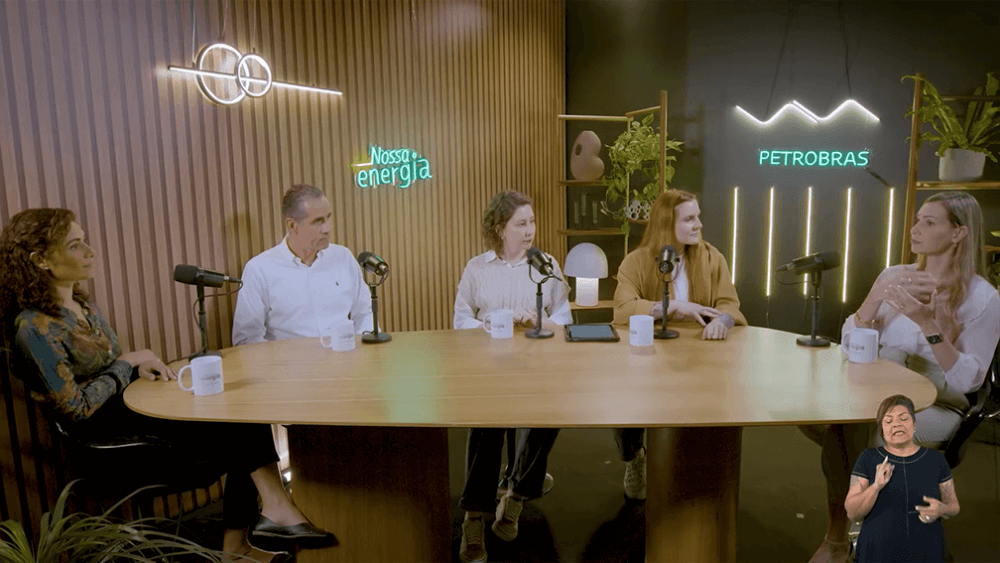 Quatro mulheres e um homem sentados lado a lado a uma mesa de madeira, em frente a letreiros neon com escritas “Nossa Energia” e “Petrobras”.