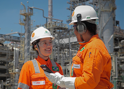 Duas mulheres com uniforme laranja e capacete branco da Petrobras conversam em frente a uma refinaria.