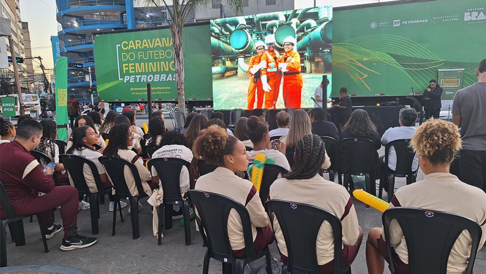 Diversas mulheres jovens sentadas em cadeiras plásticas ao ar live em frente a um palco da Caravana de Futebol Feminino Petrobras.