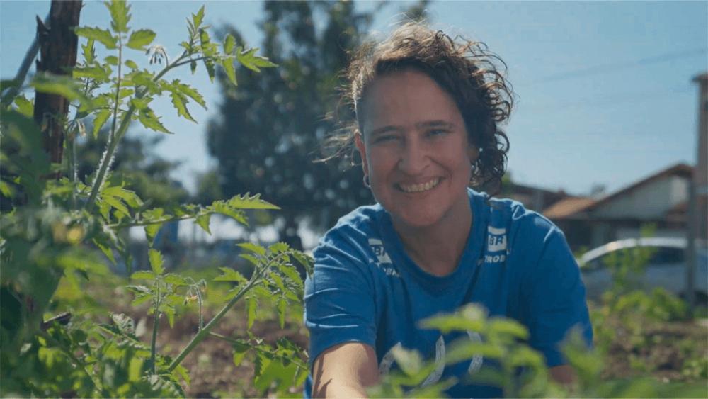 Mulher, com camisa do Projeto Corredor Caipira, sorri para a câmera em meio às folhagens de espécie nativa da Mata Atlântica, replantada pelo Projeto Corredor Caipira.