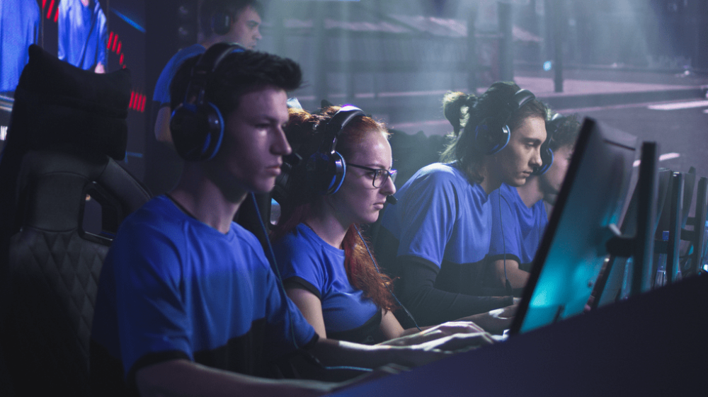 Uma mulher e três homens sentados em frente a computadores, com uniforme azul e headphones.