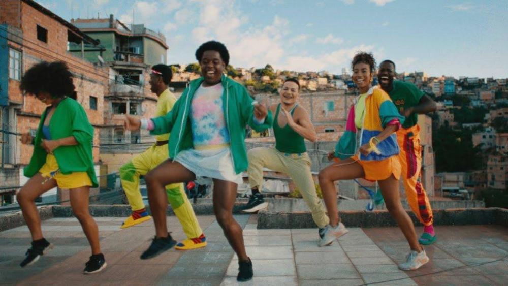 Grupo de jovens dança ao ar livre em um terraço urbano, com roupas coloridas e prédios ao fundo.
