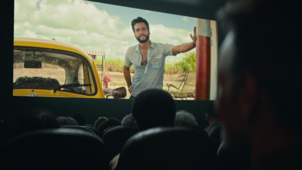 Pessoas assistem a um filme em uma sala de cinema; na tela, um homem está encostado em um carro amarelo em um cenário rural.