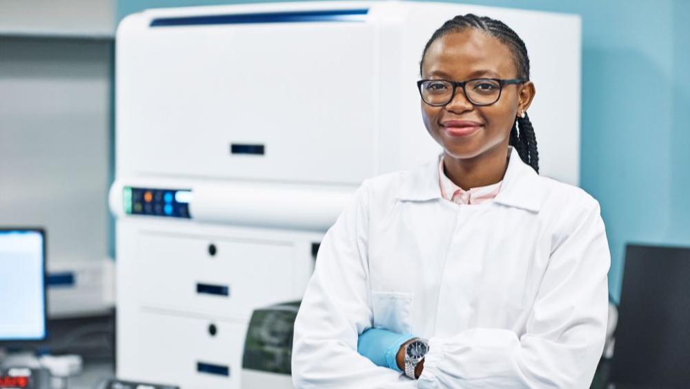Mulher negra de braços cruzados em um laboratório, usando óculos e um jaleco branco.