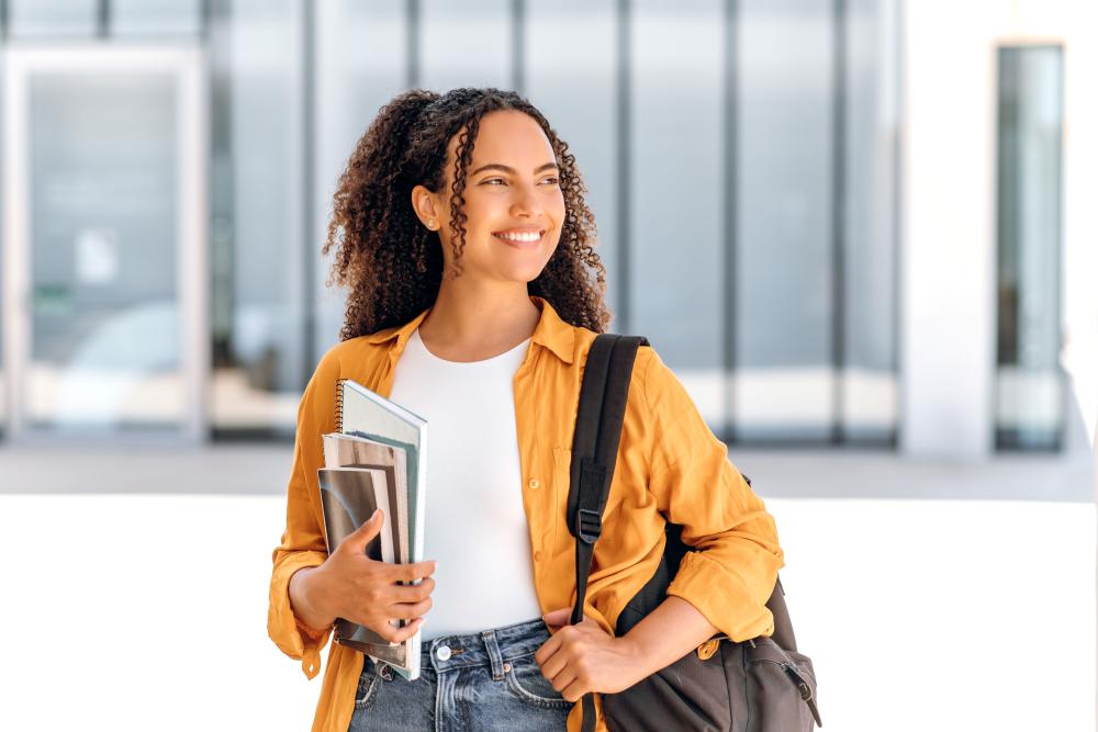 Mulher com blusa blusa laranja, calça jeans, carregando uma mochila no ombro esquerdo e livros e cadernos na mão direita