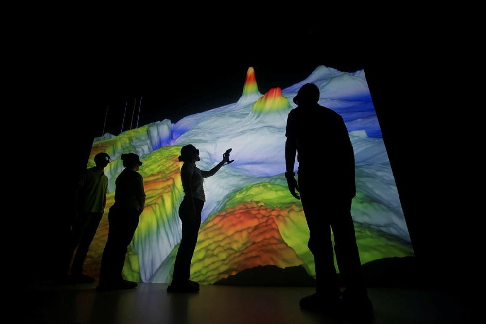 SIlhuetas de dois homens e duas mulheres em uma sala escura, em frente a uma projeção 3D colorida na parede. Todos usam óculos de realidade virtual.